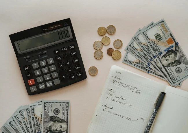 calculator, cash, coins, notebook, and pen on a desk